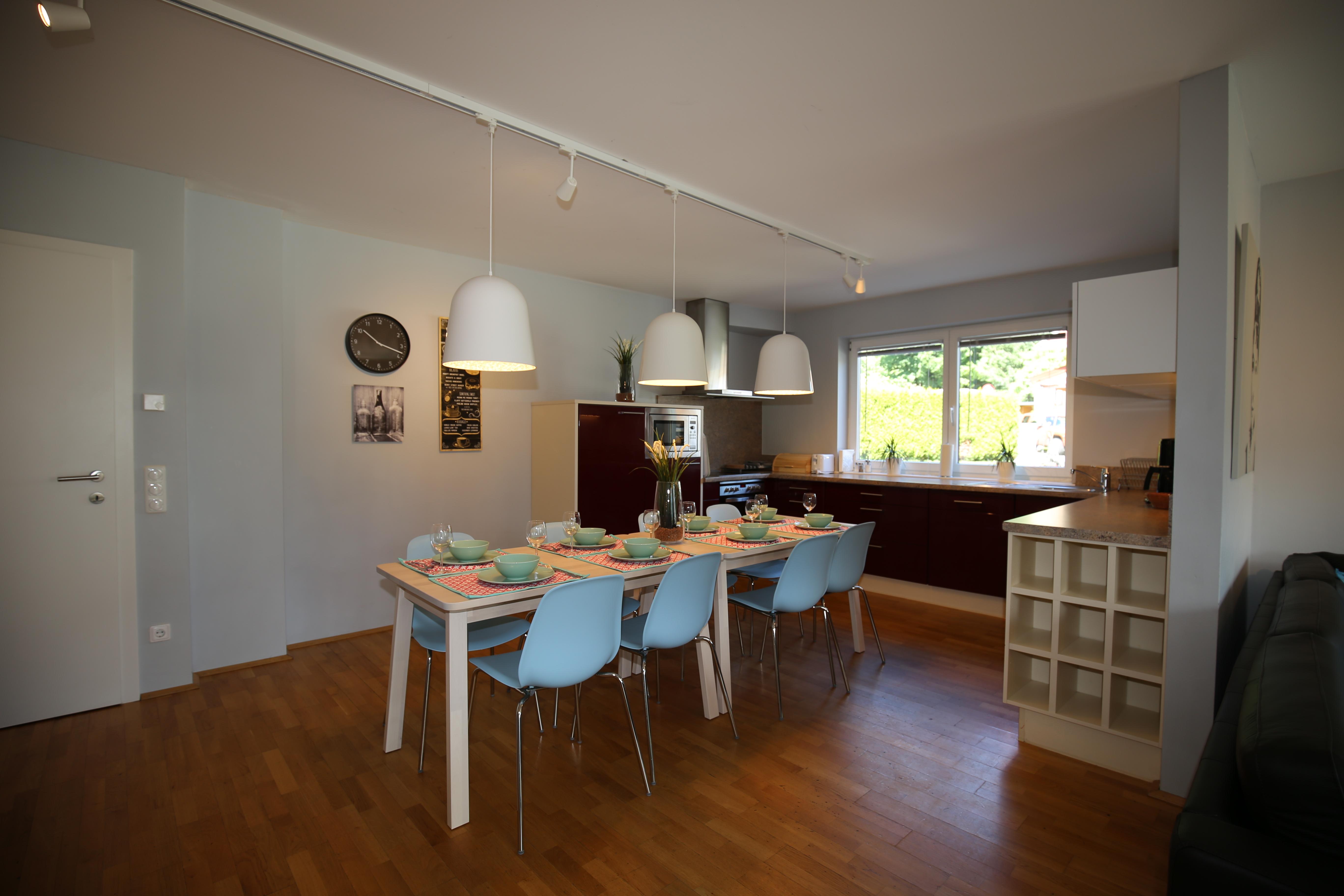 A modern kitchen with a large dining table and light blue chairs. The room has plenty of natural light and a friendly atmosphere.