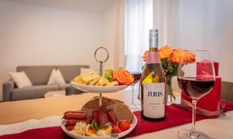 An elegant arrangement with a bottle of wine, glasses, and a selection of cheese and sausage on a table. In the background, a cozy living room with a couch can be seen.