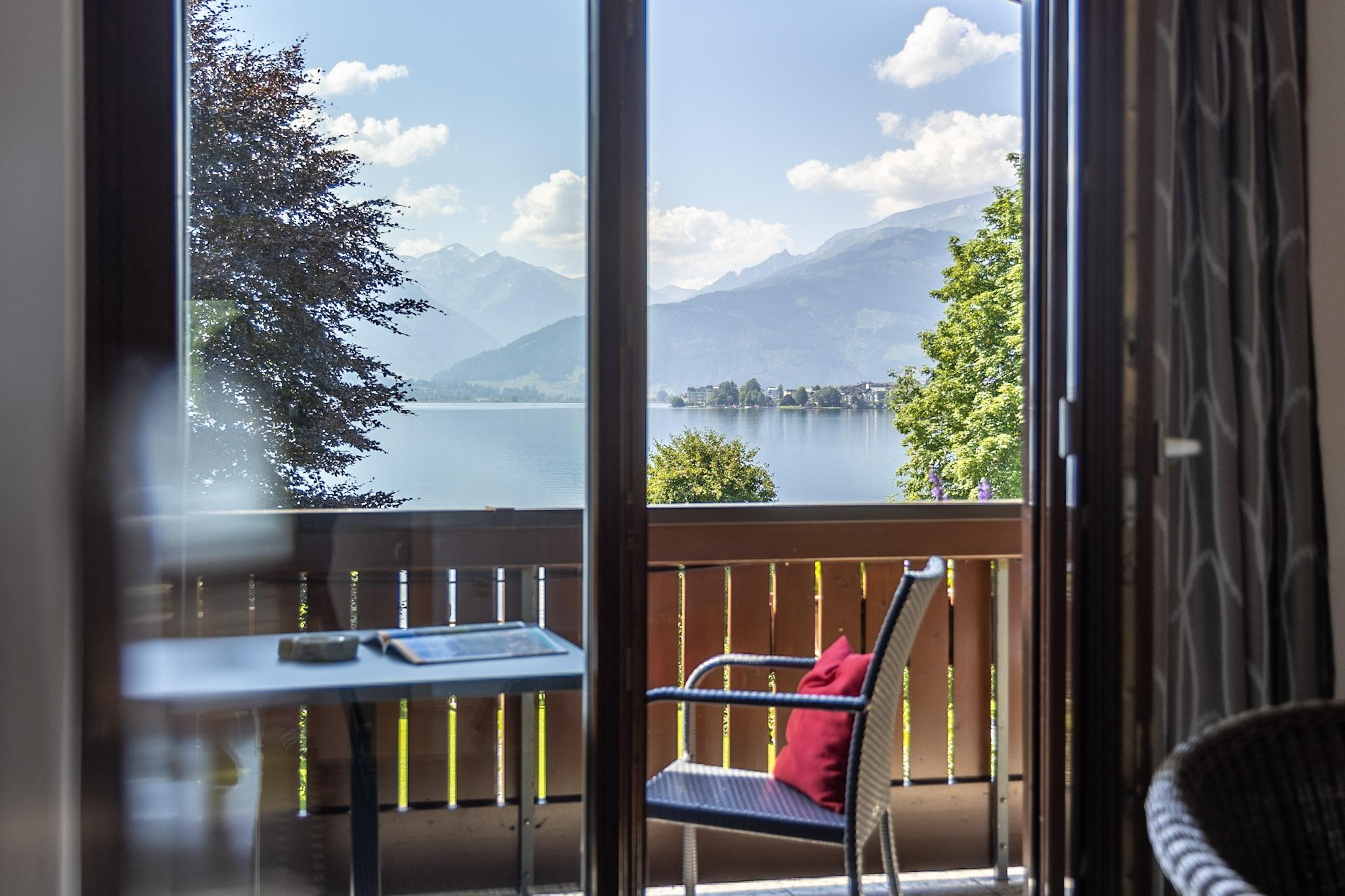 A view from a balcony over a picturesque lake with mountains in the background. On the balcony, there is a table and a chair with a red cushion.