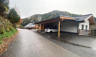 A modern house on a quiet street, surrounded by hills and trees. The parking space is occupied by a car.