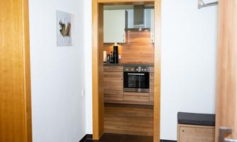 A modern kitchen with wooden cabinets and a cooking area is visible through a door. The entrance area has light walls and a seating area.