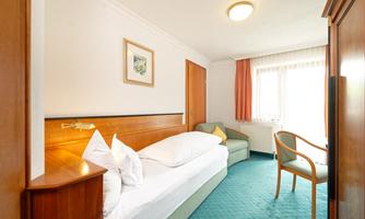 A cozy hotel room with a single bed and an armchair. The window lets in a lot of daylight and there is a small table.