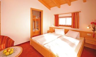 A cozy bedroom with a large double bed and a wooden ceiling beam. Next to the bed stands a table with a bowl full of apples.