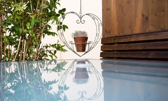 An oval flower stand in heart shape with a flower pot is in front of a wooden wall. Panels with reflective water create the foreground.