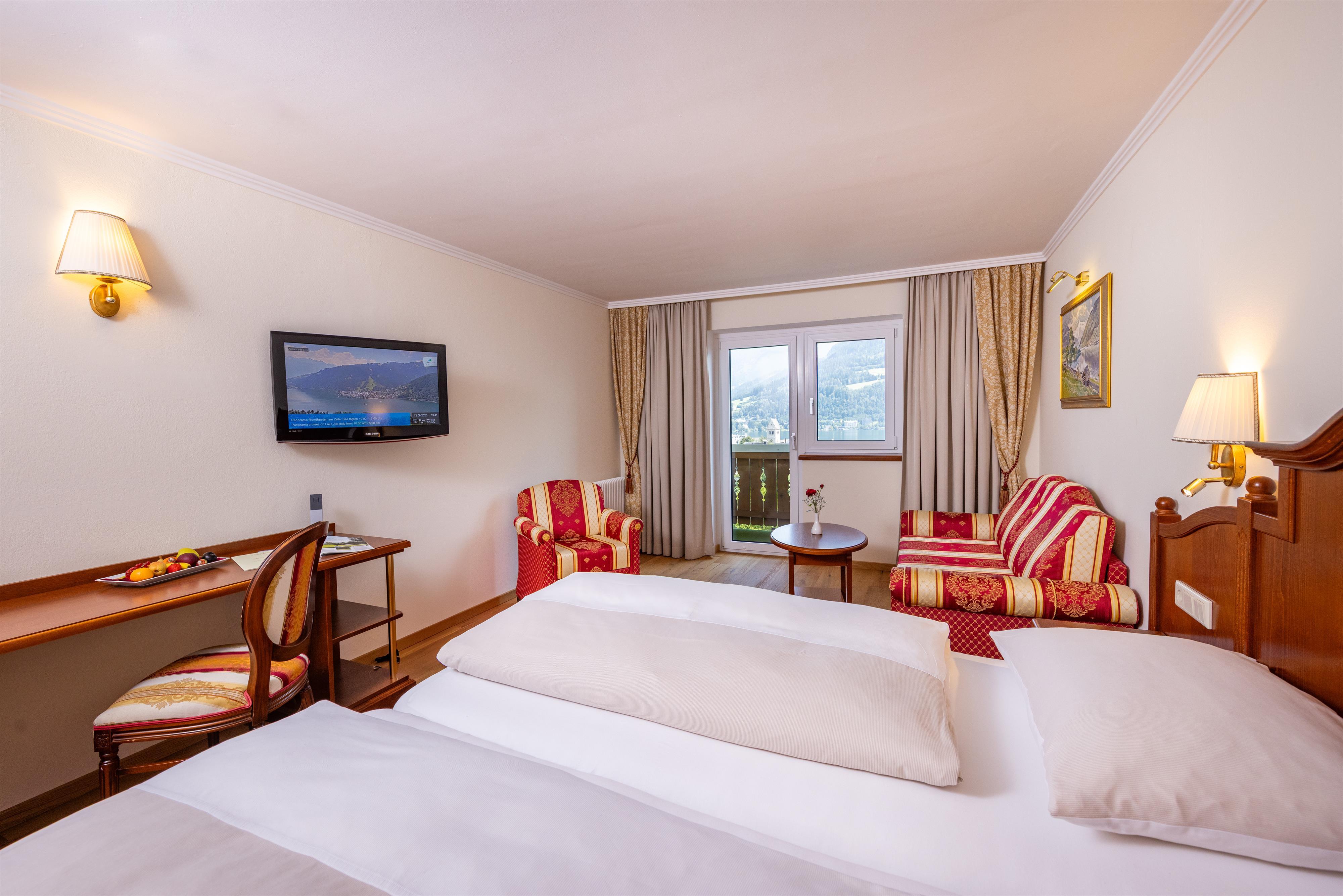 A cozy hotel room with a double bed and two red armchairs. Much daylight enters through the windows and offers a view outside.