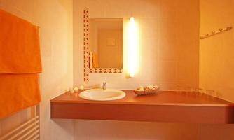 A modern bathroom with warm colors. It features a sink, a mirror, and orange towels.