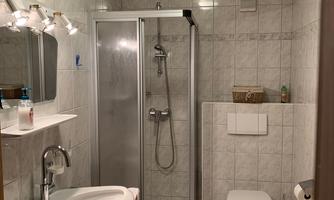 A modern bathroom with a shower cabin, a sink, and a toilet. The walls are tiled and there is bright lighting.