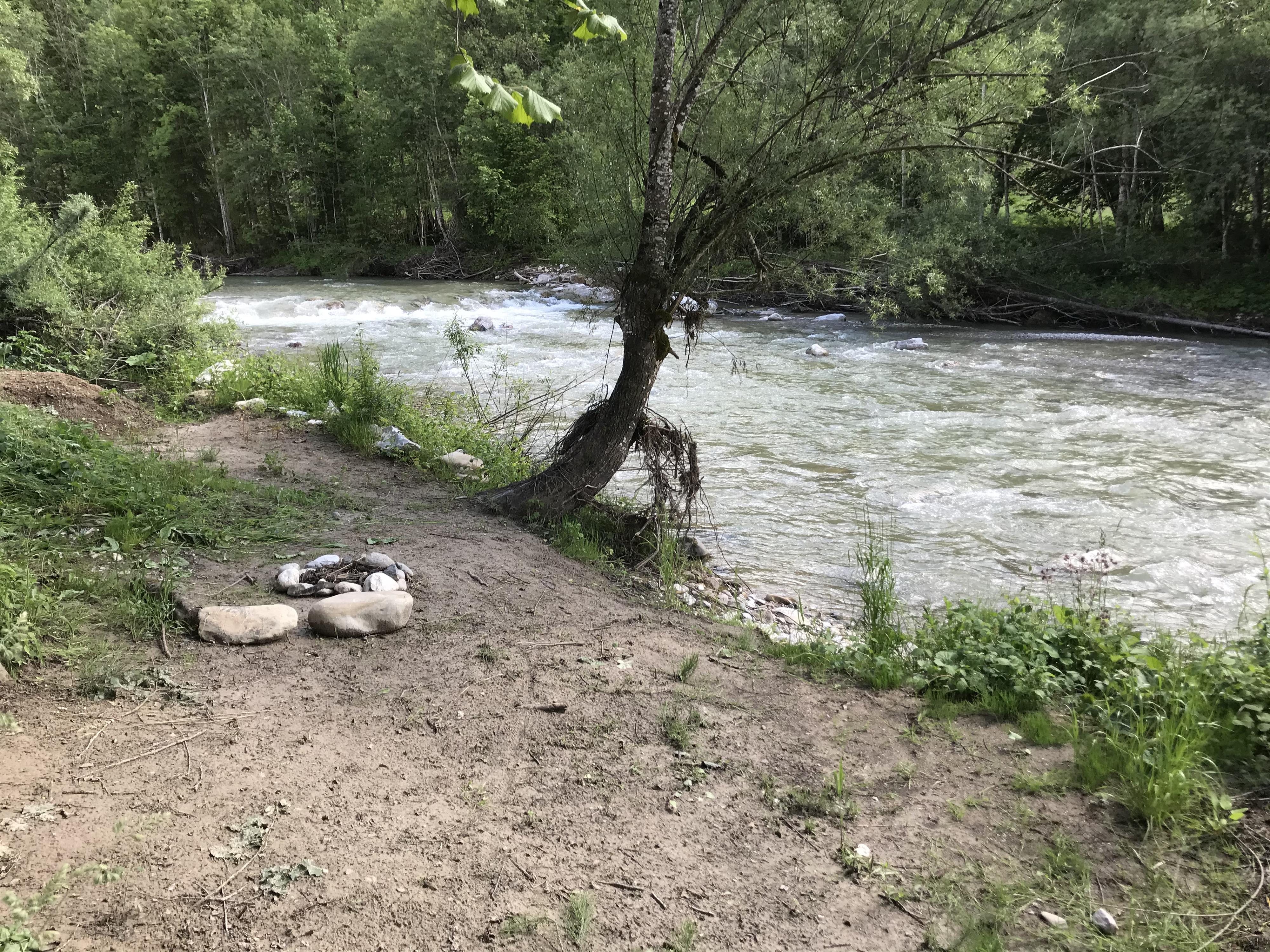 A calm river flows through a green landscape. There are some stones on the bank and there is plenty of vegetation.