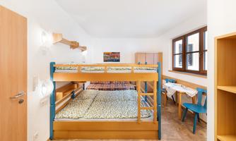 A bright room with a bunk bed and colorful bedding. On the right side, there is a table and chairs, and there are large windows that let in plenty of daylight.