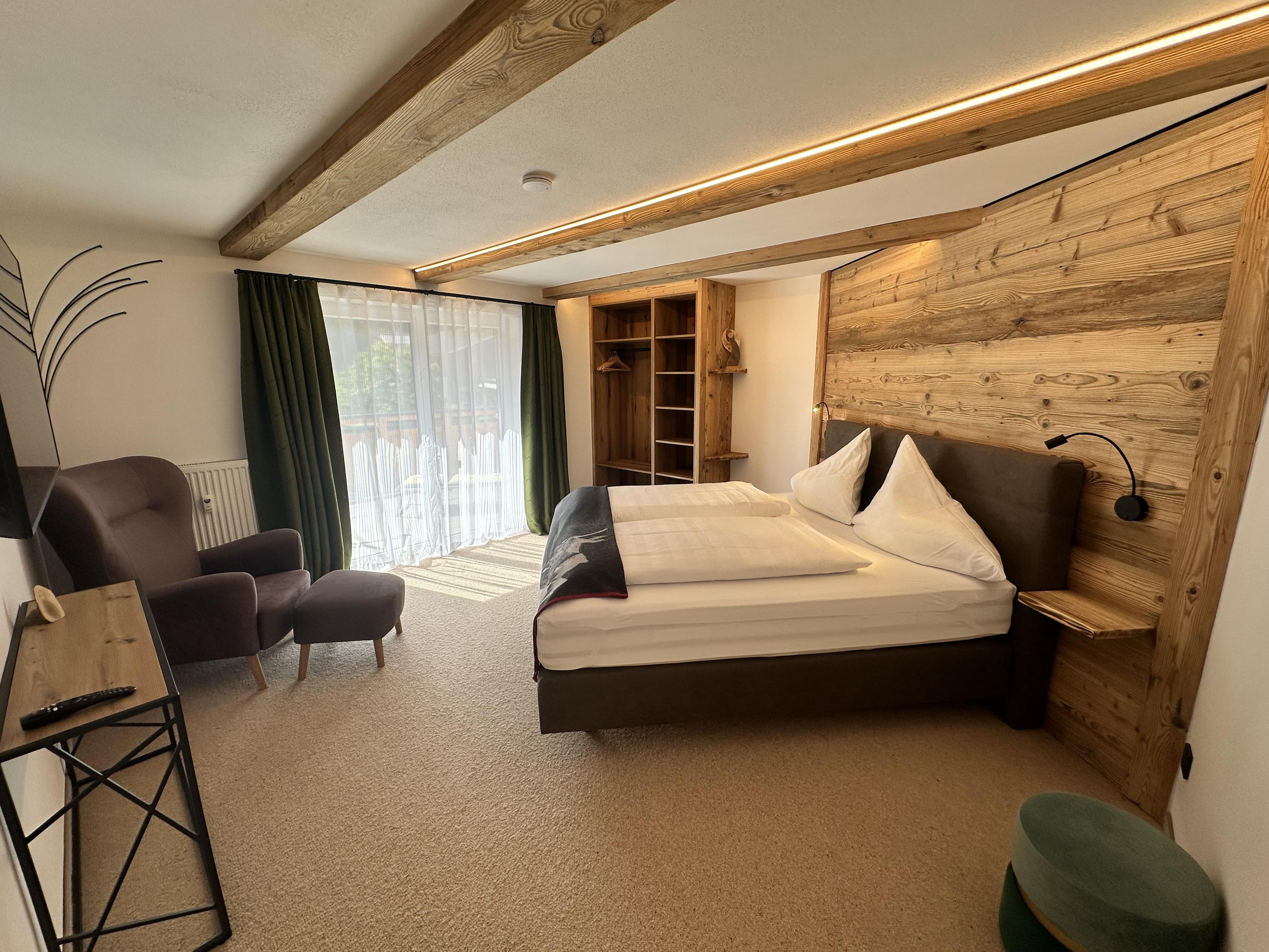 A modern room with a double bed and wooden elements. There is a comfortable armchair and large windows that let in plenty of daylight.