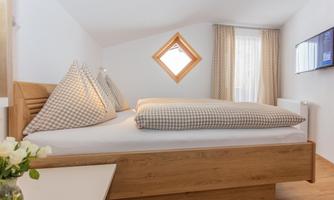 A cozy bedroom with a double bed and checkered pillows. The room has a large window and a modern TV system.