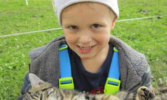 A happy boy is holding a relaxed cat in his arms. In the background, a green meadow and trees can be seen.