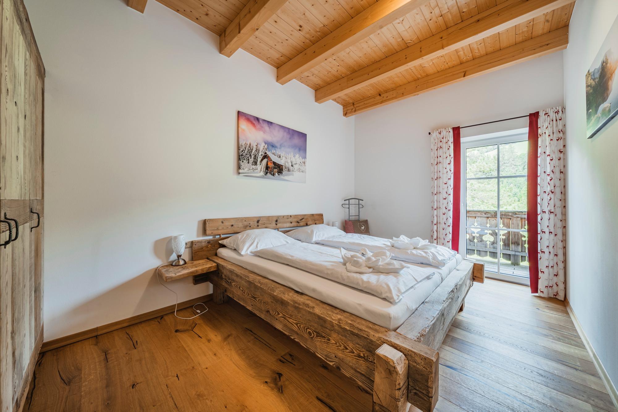 A cozy bedroom with a double bed and wooden decor. Large windows offer a view outside and let in plenty of light.