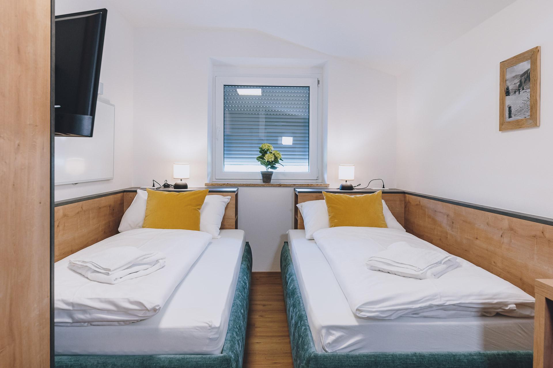 A modern guest room with two single beds and a bright decor. On the windowsill, there is a small bouquet of flowers.