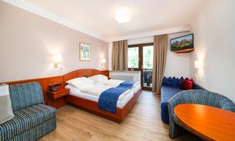 A comfortable hotel room with a double bed, a seating area, and a table. Large windows offer views of the balcony and plenty of daylight.