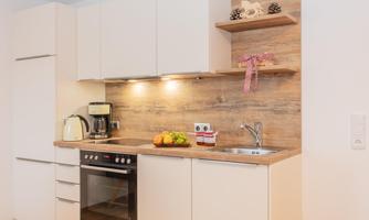 A modern kitchen with white cabinets and a wooden countertop. On the work surface are a kettle, a coffee machine, and fresh fruit.