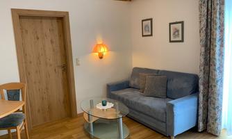 A cozy living room with a blue sofa, a glass table, and a dining table. On the wall, there is a warm light and some pictures.