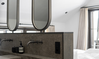 A modern bathroom with a heavy, square sink and two oval mirrors. The decor is minimalist and elegant, with natural tones.