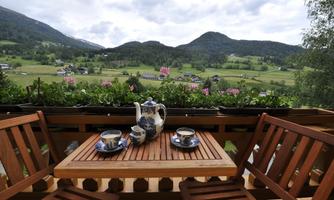 A cozy balcony with a wooden table and two chairs. On the table are a teapot and cups, overlooking a green landscape and mountains.