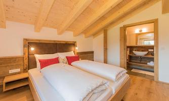 A cozy bedroom with a wooden ceiling and two beds. In the background, a modern bathroom is visible.