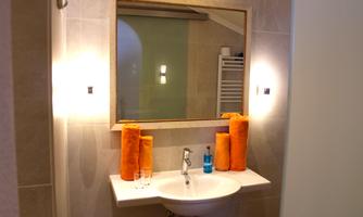 A modern bathroom with a sink and a large mirror. On the shelf are orange towels and a bottle with blue contents.