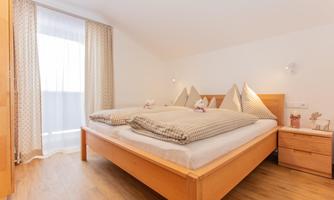 A cozy bedroom with a large double bed and wooden details. Bright curtains let in plenty of daylight.