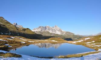 A quiet mountain lake surrounded by snow-covered meadows and majestic mountains. The clear sky reflects in the water, creating a peaceful atmosphere.
