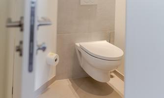 A modern bathroom with a white toilet and a gray tiled floor. Next to the toilet is a roll of toilet paper.