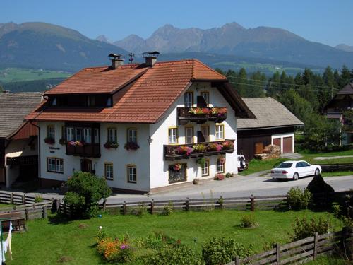 A cozy house with blooming balconies in a hilly landscape. In the background, majestic mountains and a green meadow can be seen.