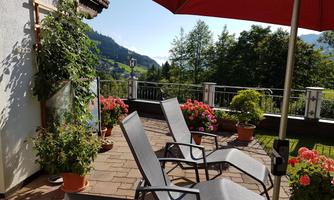 A sunny terrace with deck chairs and blooming plants. In the background, the green mountains are visible.