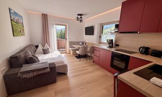 A modern living kitchen with red kitchen cabinets and a cozy living area. Large windows offer a view of nature.