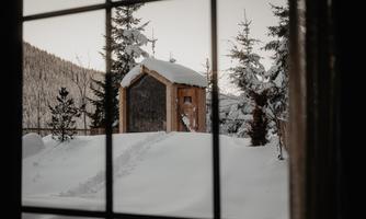 A snowy winter landscape with a small wooden house. Trees frame the scene and the footprints in the snow suggest recent movement.