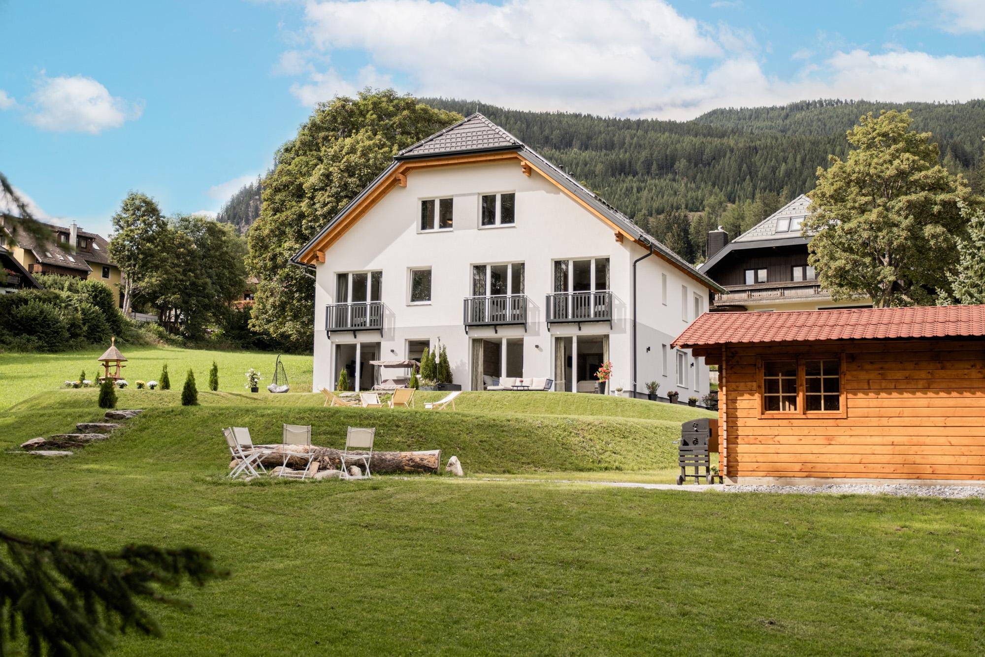 A modern house in a green, open garden. In the background, gentle hills and trees can be seen.