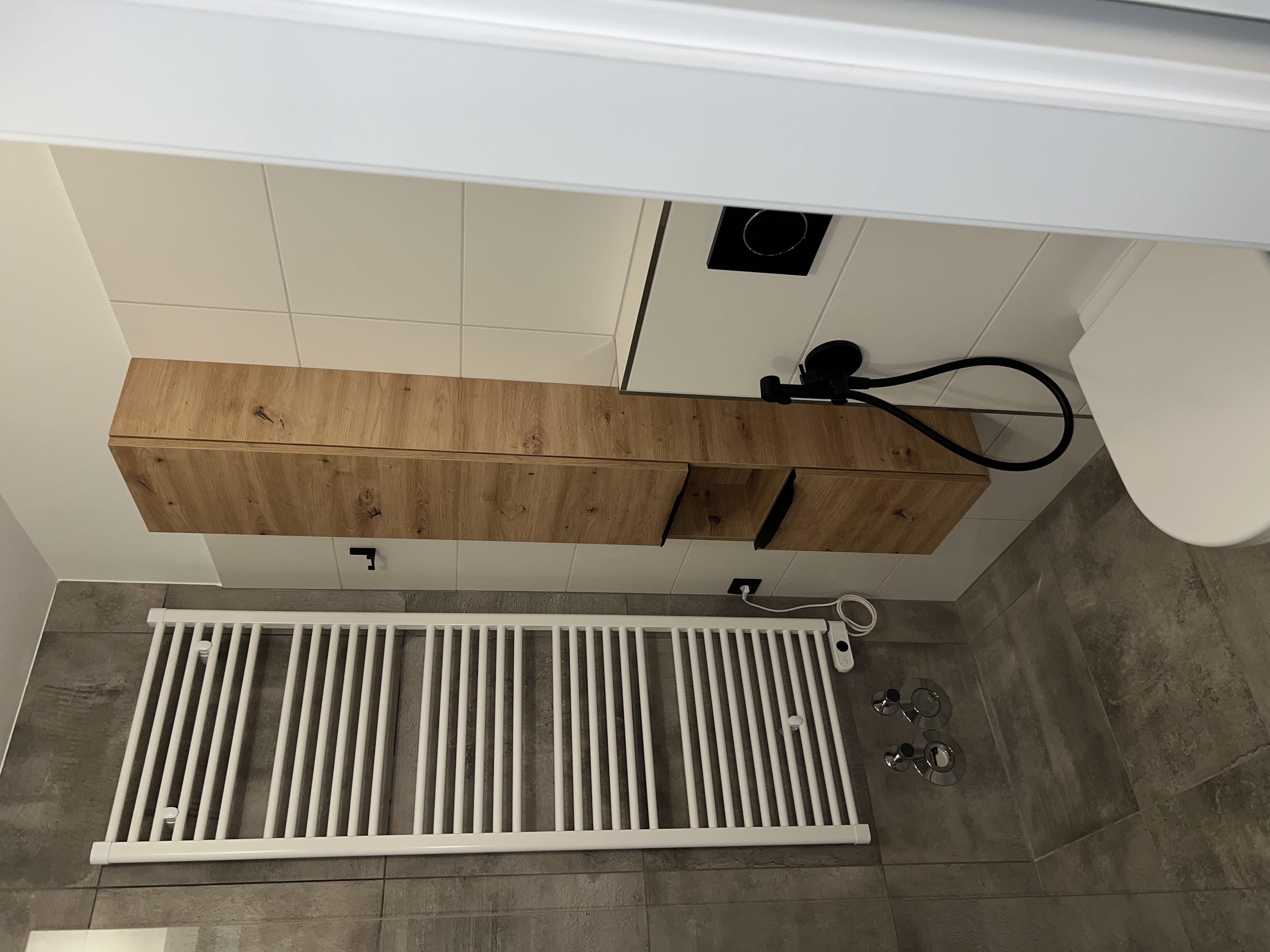 A modern bathroom with a shower, a white towel warmer, and a wooden cabinet. The walls are light tiled, and the floor is grey.