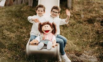 Two boys are sitting on a slide and smiling at the camera. Between them stands a doll that looks cheerful.