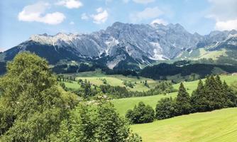 A picturesque mountain landscape with green meadows and dense forests. In the background, majestic peaks are visible under a clear blue sky.