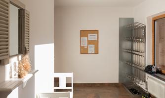 A bright entrance area with a seating area and a notice board. Shoe shelves are against the wall, and sunlight is streaming through the window.