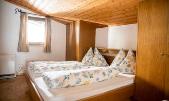 A cozy bedroom with two single beds and wooden ceilings. The walls are bright, and there are large windows with curtains.
