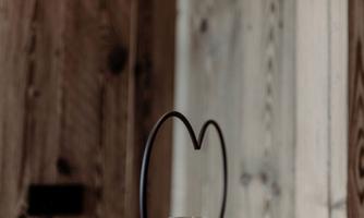 A stylish heart-shaped candle holder is placed on a wooden table. Next to it is a decorative glass containing a small plant.