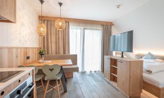 A modern apartment with wooden interiors and large windows. There is a dining table, chairs, and a television in the room.