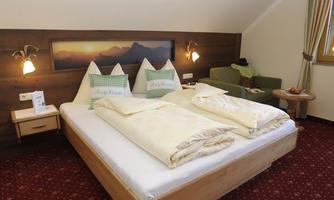 A cozy bedroom with a large bed and two pillows. In the background, there is a picture of mountains and a seating area next to the window.
