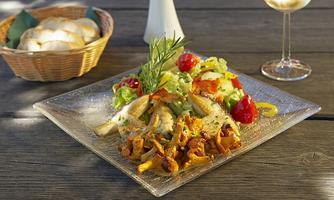 A plate with fried mushrooms, vegetables, and salad. In the background, there is a basket with rolls and a glass of white wine.