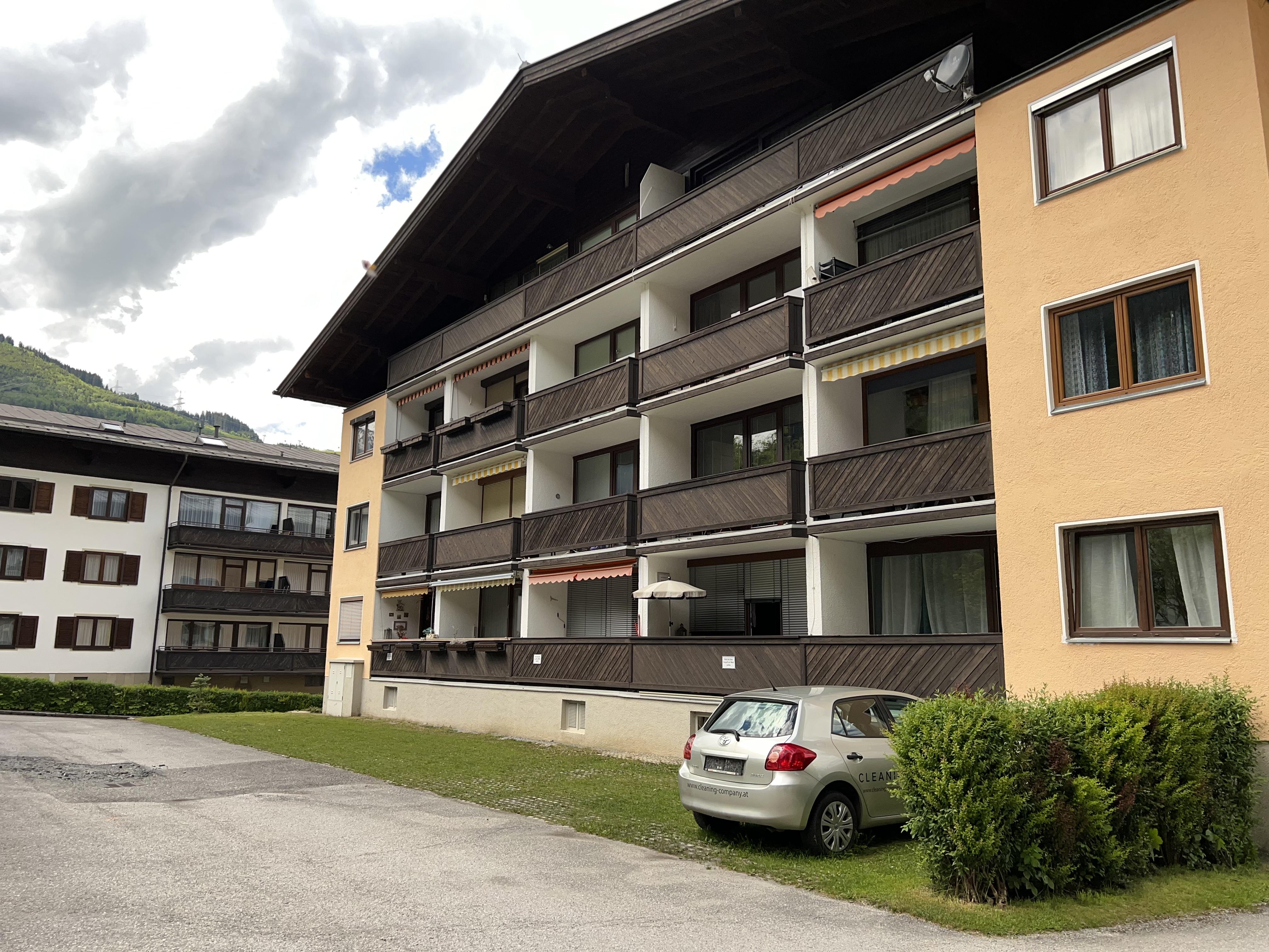 A residential complex with several floors and balconies. In the foreground, there is a gray car on a green meadow.