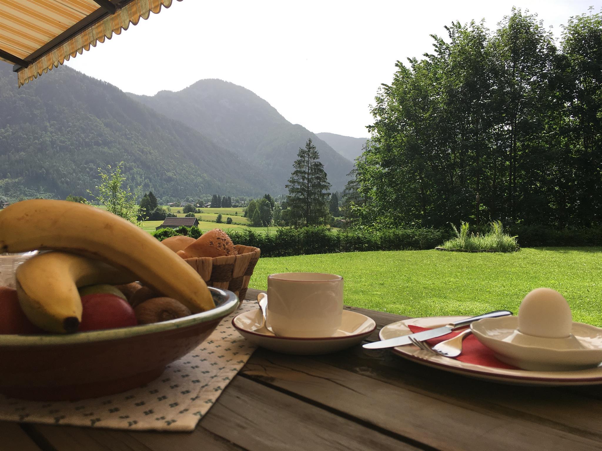 A table with fresh fruit, coffee, and a boiled egg. In the background, you can see the mountains and a green meadow.