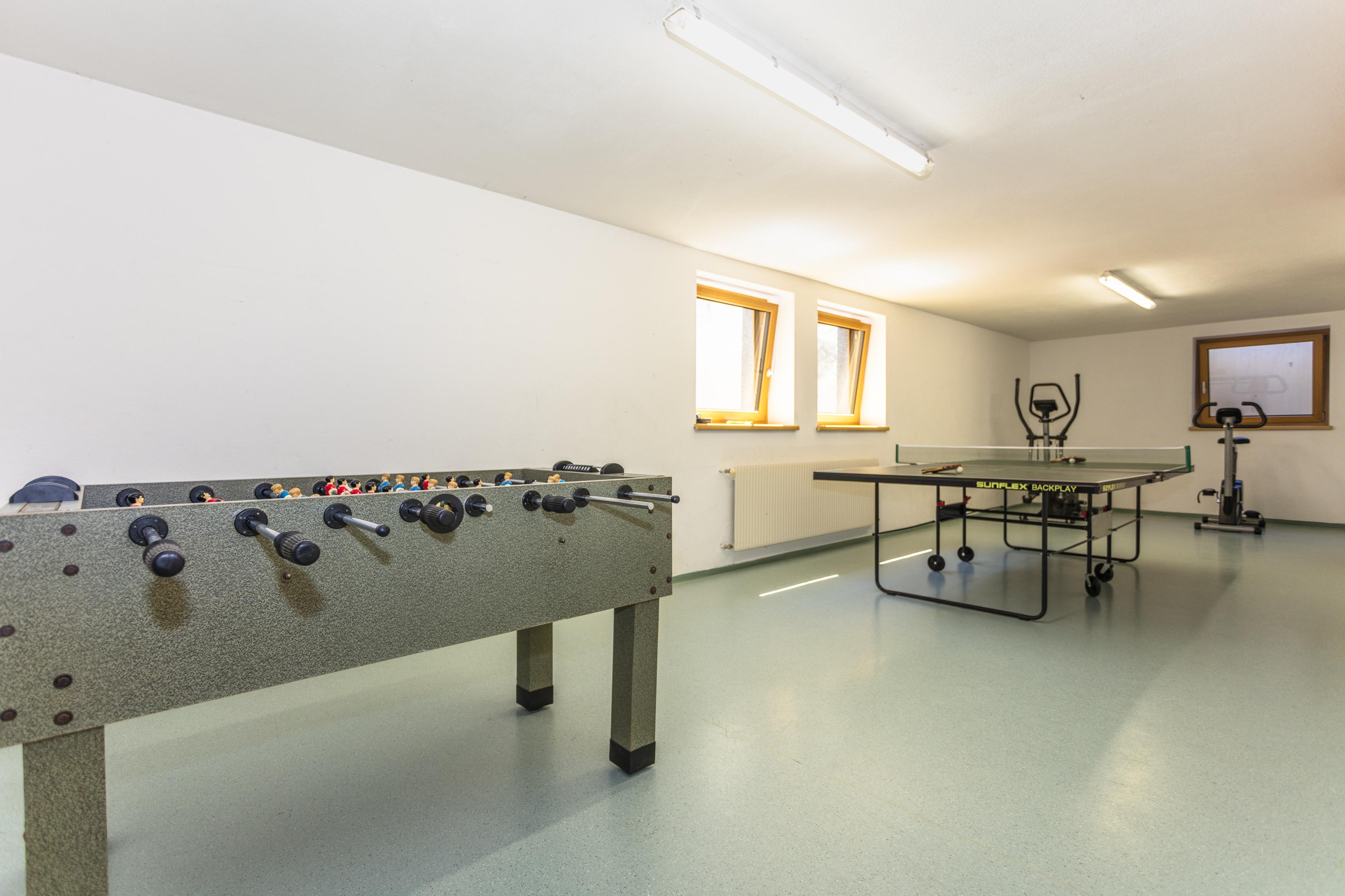 A room with a foosball table and a billiard table. The walls are light and there are windows that provide natural light.