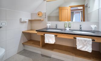A modern bathroom with two sinks and wooden shelves. The room has bright tiles and a large window that provides good lighting.