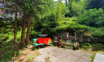 A cozy outdoor area with a bench and red cushions, surrounded by trees and green vegetation. The paved floor and decorative elements create an inviting atmosphere.