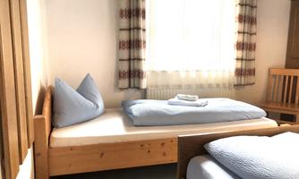 A simple bedroom with two wooden beds. The room has a window with curtains and a chair.
