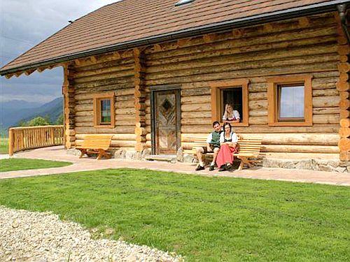 A beautiful wooden house with a green lawn and benches in front. A family relaxes on the porch.