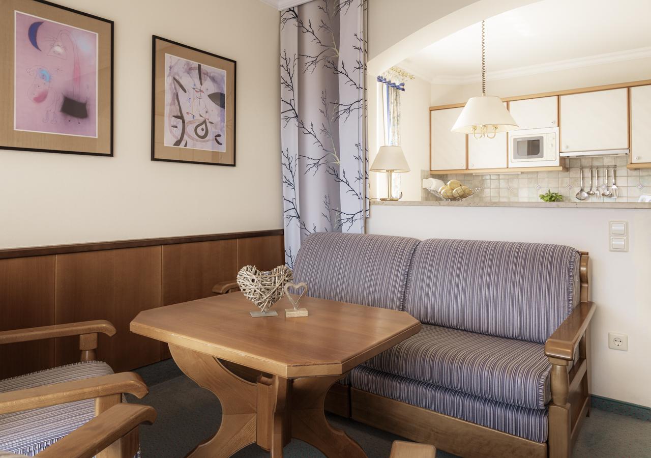 A cozy living room with a wooden table and a blue striped sofa. In the background, there are pictures on the wall and a small kitchenette visible.